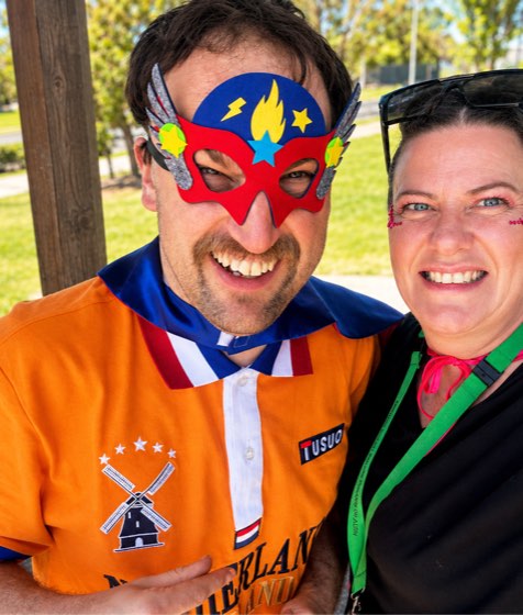 A participant and his support worker wearing superhero masks at a community day, both laughing.