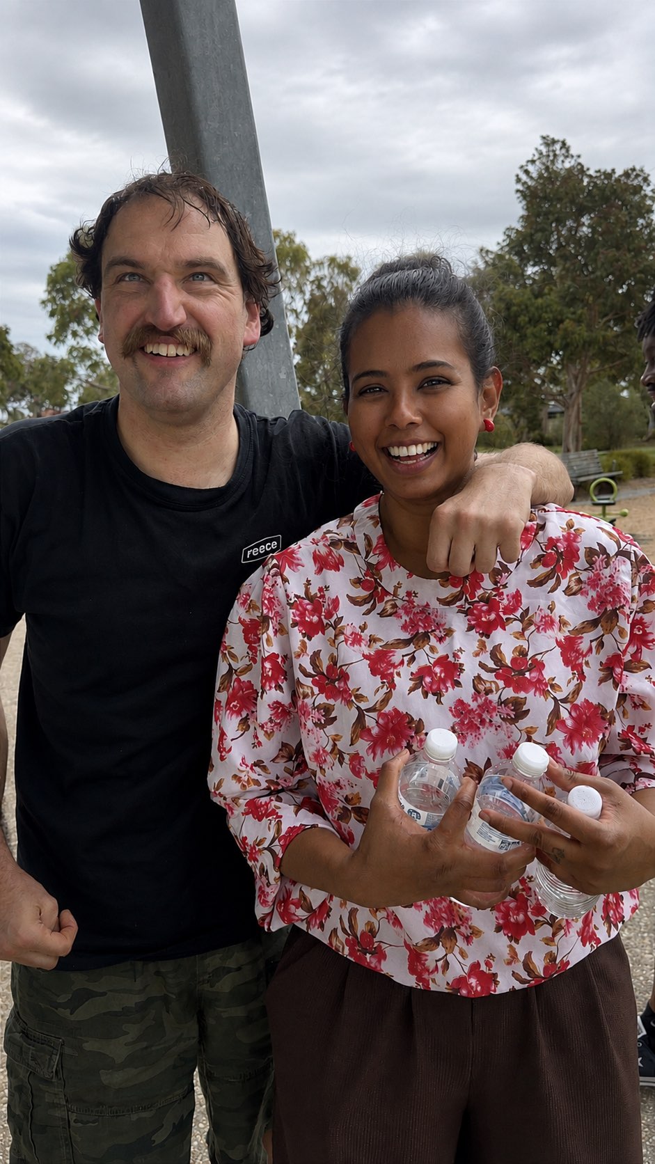 A support worker and a participant standing arm in arm, laughing together at a local park.